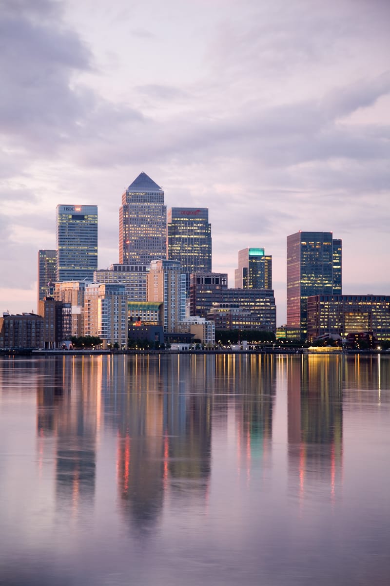 The Canary Wharf cluster reflected in calm water at sunrise under a pastel sky